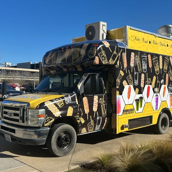 A Bubblebee food truck, black and yellow with illustrations of boba drinks and corn dogs, parked outdoors.