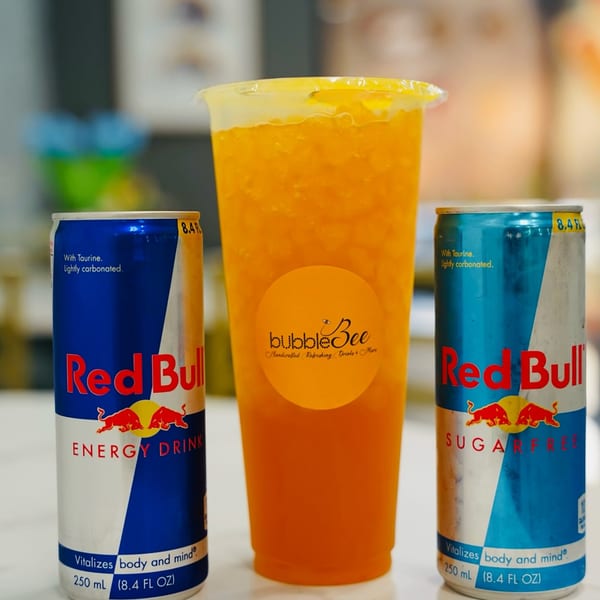 A BubbleBee mango drink in a clear cup, flanked by two Red Bull cans on a white surface.