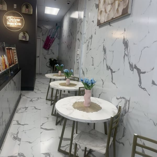 A narrow dining area with marble-patterned tables, chairs, and walls, featuring blue flowers and a neon sign.