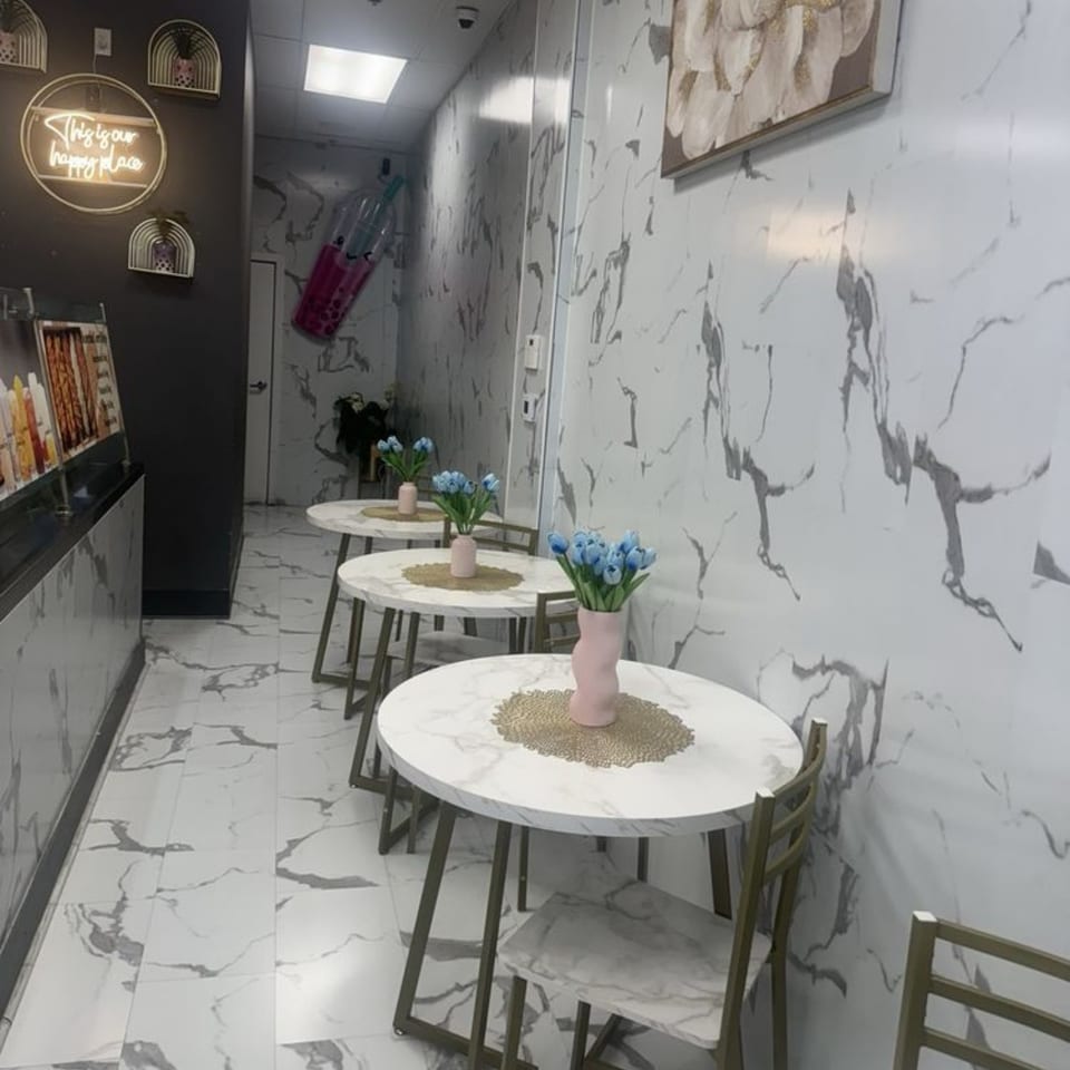 A narrow dining area with marble-patterned tables, chairs, and walls, featuring blue flowers and a neon sign.