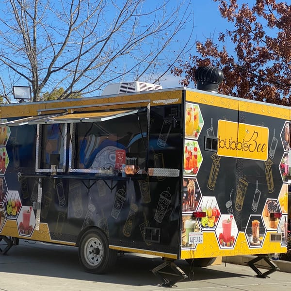 Black and yellow Bubble Bee food truck with drink images, parked under a blue sky and trees.